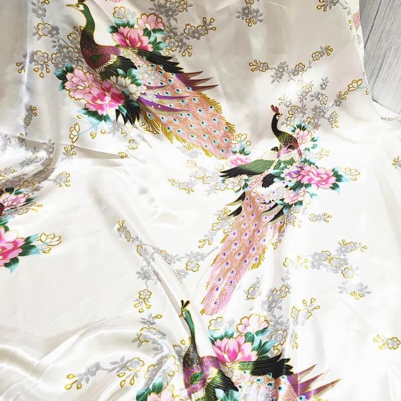 Le Tissu satin imprimé paon au mètre de Longan Craft France est un tissu satin de couleur claire avec des motifs de paons colorés, de fleurs roses, de feuilles vertes et de branches grises, idéal pour créer des accessoires élégants.