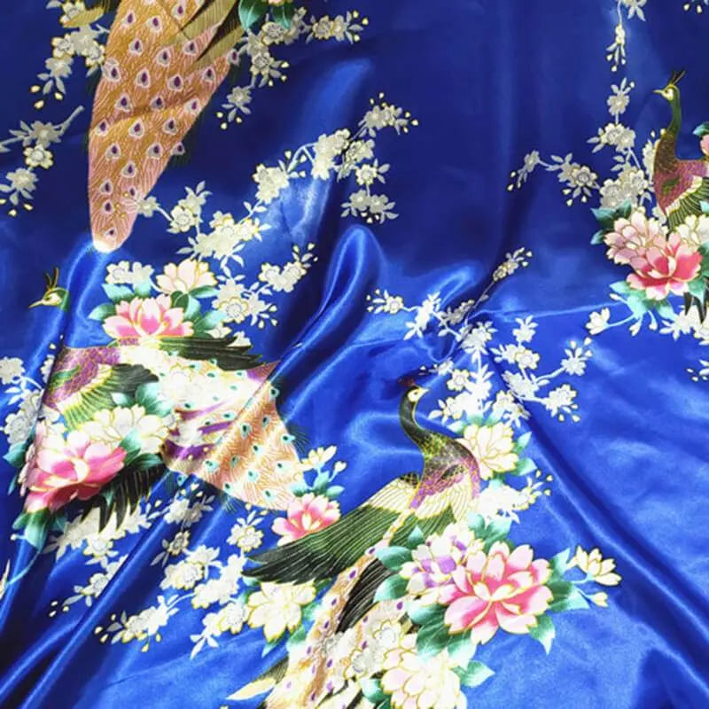Le Tissu satin imprimé paon au mètre de Longan Craft France présente des motifs colorés de paons, fleurs roses et blanches, et feuilles vertes. Sa texture satinée est idéale pour réaliser des accessoires élégants ou une décoration intérieure raffinée.