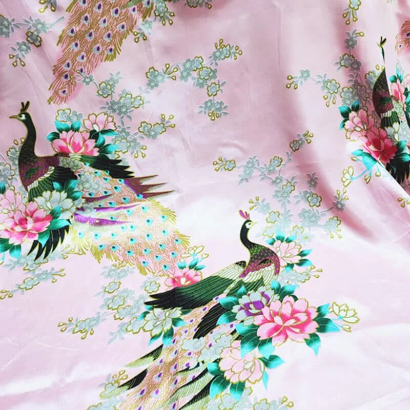 Le "Tissu satin imprimé paon au mètre" de Longan Craft France présente des paons colorés et des motifs floraux sur un satin rose, parfait pour des accessoires élégants et des projets accrocheurs.