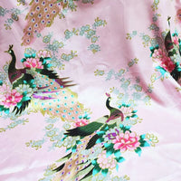 Le "Tissu satin imprimé paon au mètre" de Longan Craft France présente des paons colorés et des motifs floraux sur un satin rose, parfait pour des accessoires élégants et des projets accrocheurs.