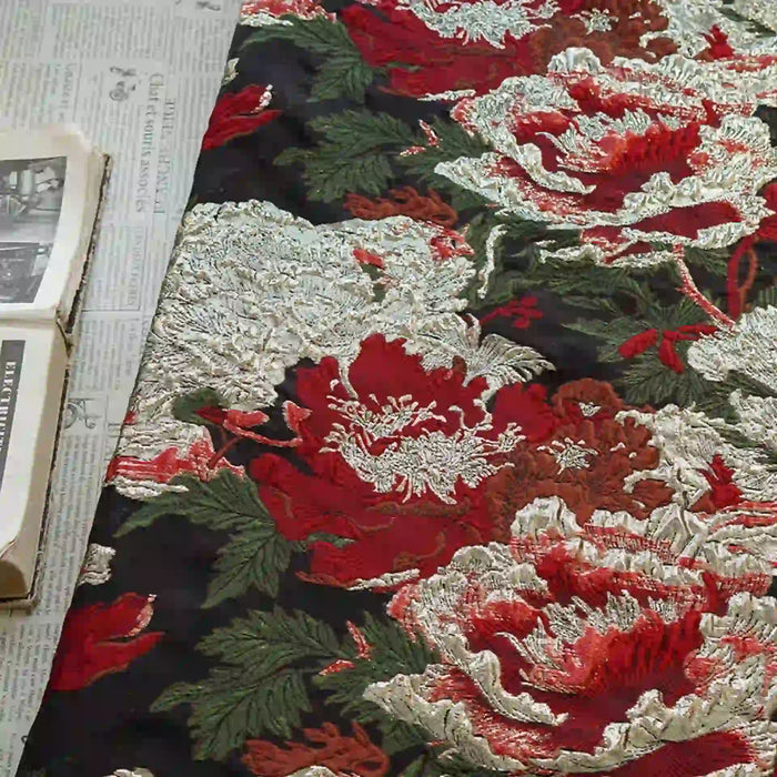 The Tissu brocart métallisé embossé pivoine de Longan Craft France, un brocart jacquard floral noir et doré de luxe pour robes de soirée, est présenté à côté d'un journal ouvert et d'un livre aux photos en noir et blanc.