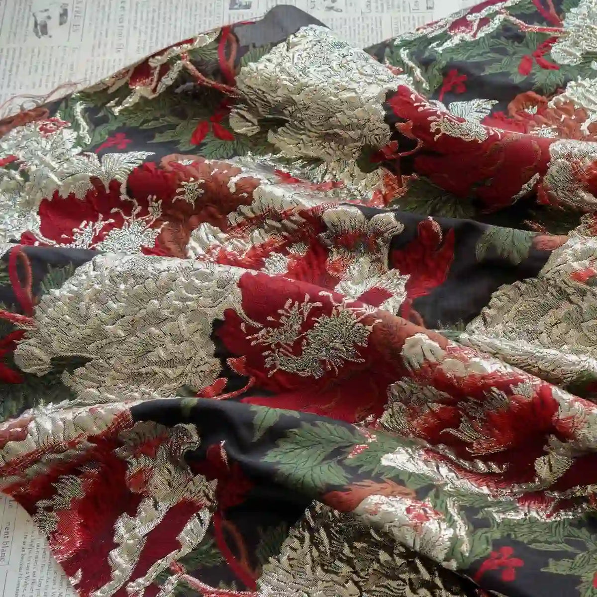 Gros plan sur le Tissu brocart métallisé embossé pivoine de Longan Craft France, un tissu jacquard floral noir et doré de luxe pour les robes du soir, qui présente des fleurs ornées d'or et de rouge sur un fond sombre, drapées sur un texte imprimé.