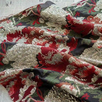 Gros plan sur le Tissu brocart métallisé embossé pivoine de Longan Craft France, un tissu jacquard floral noir et doré de luxe pour les robes du soir, qui présente des fleurs ornées d'or et de rouge sur un fond sombre, drapées sur un texte imprimé.