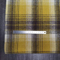 Une règle argentée est posée horizontalement sur le tissu flanelle épaisse brossée un côté à carreaux, coloré jaune, blanc et noir en polyester-coton, posé sur une surface sombre et texturée de Longan Craft France.