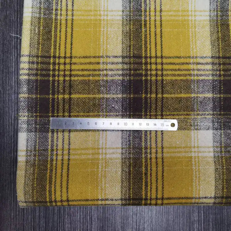 Une règle argentée est posée horizontalement sur le tissu flanelle épaisse brossée un côté à carreaux, coloré jaune, blanc et noir en polyester-coton, posé sur une surface sombre et texturée de Longan Craft France.