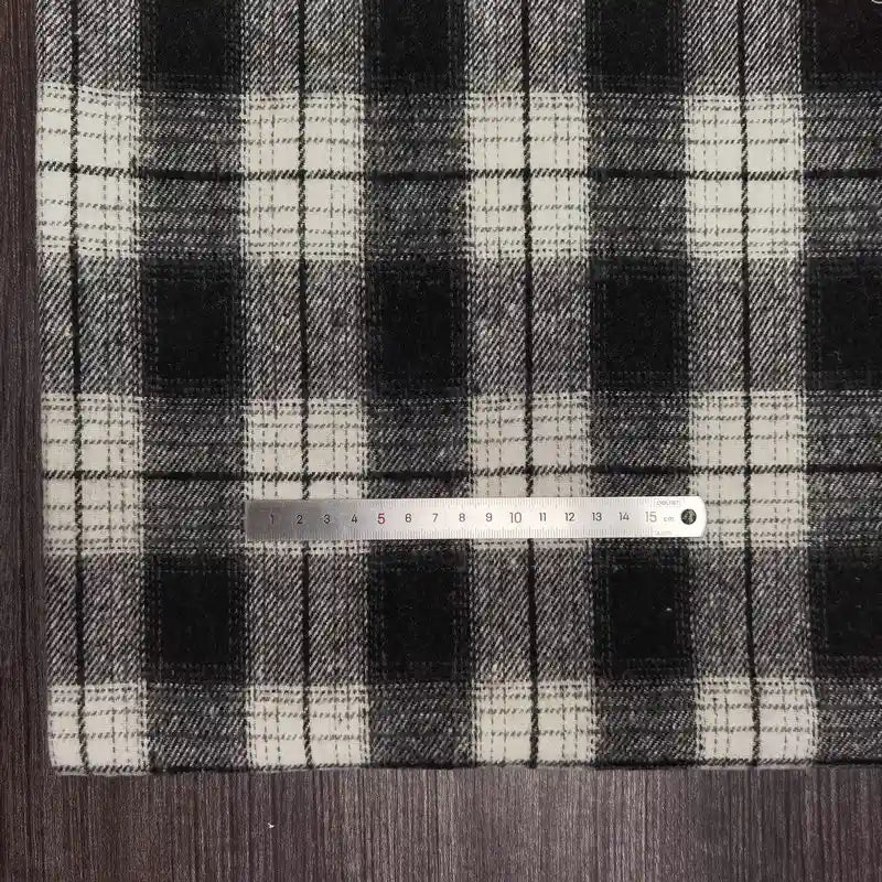 Une règle métallique est posée horizontalement sur le tissu flanelle épaisse brossée un côté à carreaux de Longan Craft France, un plaid confortable en polyester-coton vendu au mètre, présenté sur une surface en bois sombre.