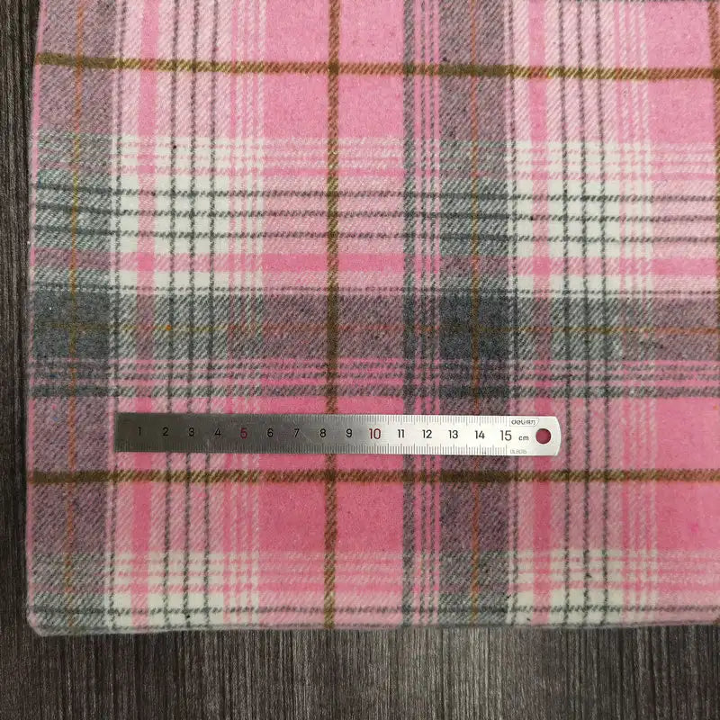 Un tissu flanelle épaisse brossée un côté à carreaux rose, blanc et gris de Longan Craft France est présenté sur une surface en bois sombre avec une règle métallique d'environ 16 cm pour l'échelle. Parfait pour le confort de l'automne, ce tissu en polyester-coton est vendu au mètre.