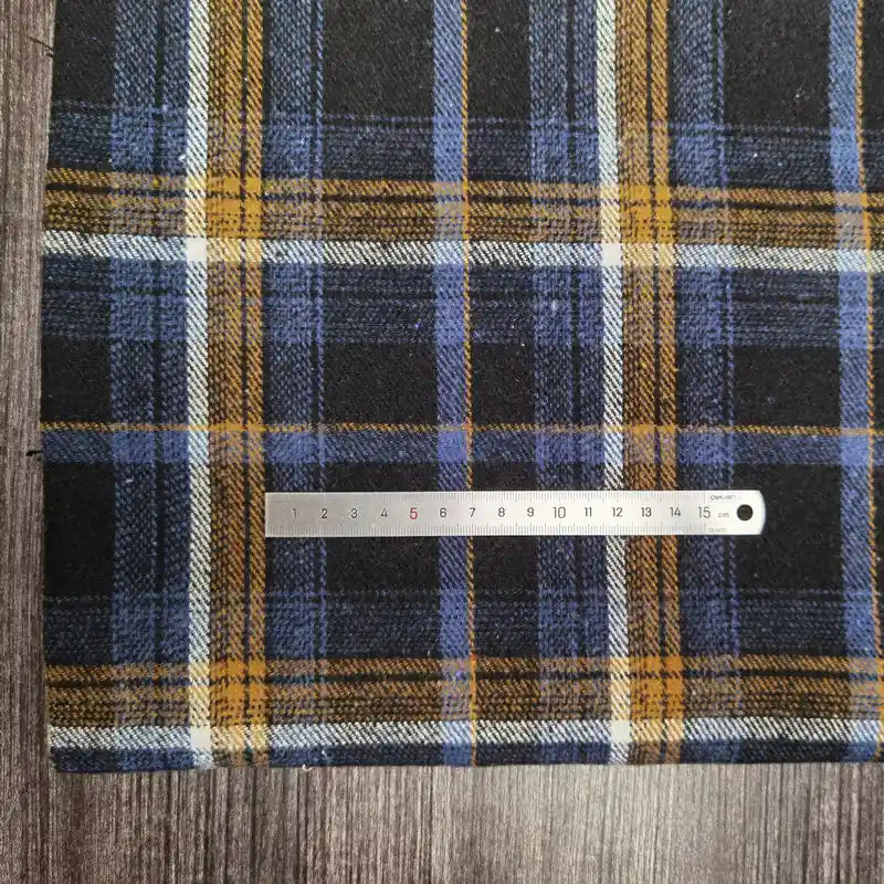 A piece of Tissu flanelle épaisse brossée un côté à carreaux Longan Craft France, coloré bleu, jaune et blanc en polyester-coton, est posé sur du bois. Une règle métallique graduée en centimètres traverse le tissu.