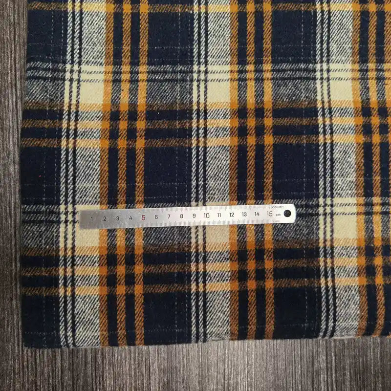 Une règle en métal est posée sur le tissu flanelle épaisse brossée un côté à carreaux (polyester-coton) de Longan Craft France, à carreaux jaune, marine et blanc. La règle mesure environ 16 cm. Le tissu est posé sur une surface grise texturée.
