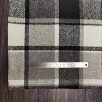 Sur un tissu flanelle épaisse brossée un côté à carreaux noir, blanc et gris de Longan Craft France, une règle métallique indique les centimètres ; le tissu coloré en polyester-coton est posé sur une surface en bois foncé.