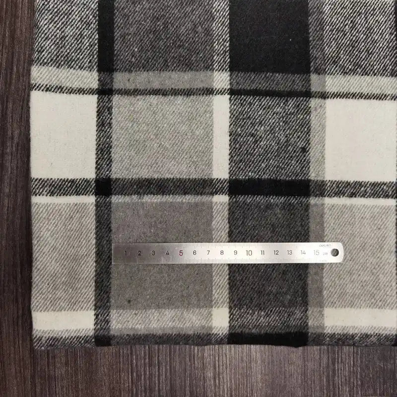 Sur un tissu flanelle épaisse brossée un côté à carreaux noir, blanc et gris de Longan Craft France, une règle métallique indique les centimètres ; le tissu coloré en polyester-coton est posé sur une surface en bois foncé.