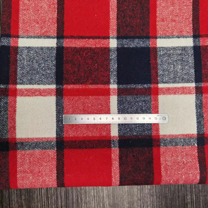 La flanelle épaisse brossée à carreaux de Longan Craft France est recouverte d'une règle en métal avec des repères en centimètres. Il s'agit d'un tissu à carreaux confortable en polyester et coton, de couleur rouge, blanche, noire et bleu marine, posé sur une surface texturée en bois.