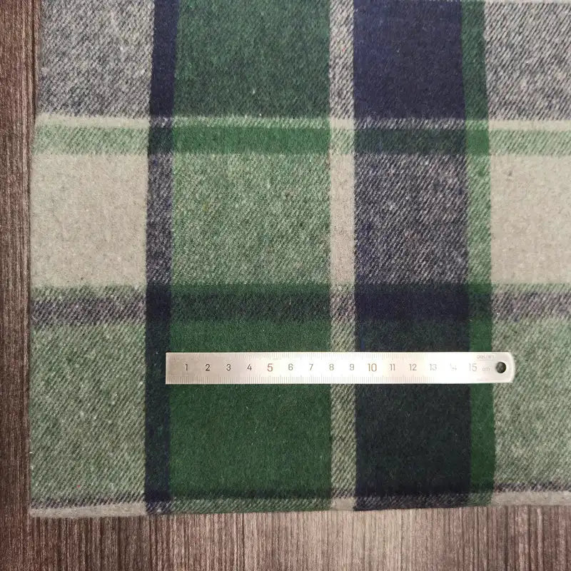 Une règle argentée en centimètres repose sur du Tissu flanelle épaisse brossée à carreaux (Longan Craft France) en plaid polyester-coton vert, marine et crème. Le doux tissu d'automne et la surface en bois se révèlent en dessous.