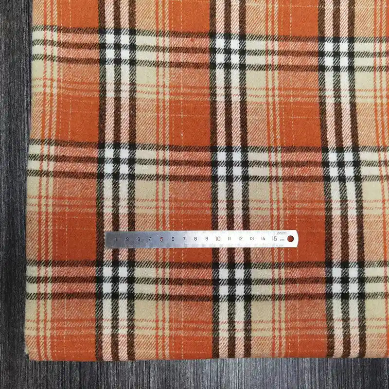 Un épais tissu écossais en flanelle brossée de Longan Craft France, dans les tons orange, beige et noir, est posé sur une surface en bois sombre. Une règle centimétrique argentée mesure son motif. Mélange idéal de polyester et de coton pour des projets d'automne douillets.