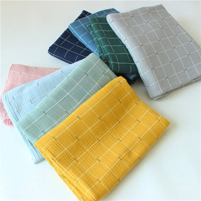 Sept pièces pliées en Tissu double gaze de coton à carreaux au mètre par Longan Craft France, en jaune, bleu clair, vert menthe, rose, bleu marine, vert et gris avec un motif quadrillé blanc, disposées sur une surface blanche.