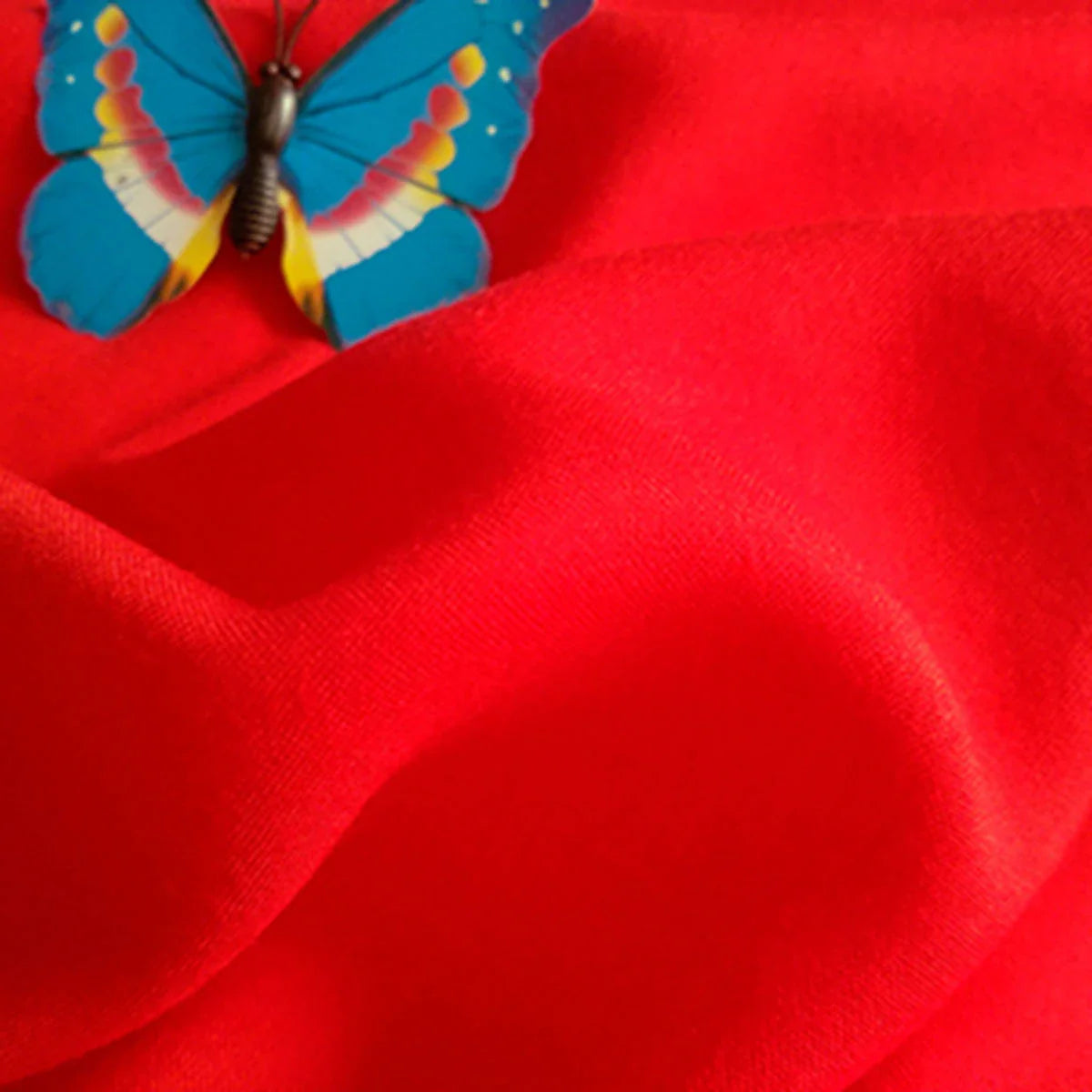 Un papillon bleu avec des marques jaunes et rouges repose sur un Tissu jersey viscose uni au mètre rouge vif de Longan Craft France, un tissu 100 % viscose rayonne idéal pour la confection de robes, dont la texture présente des plis visibles.