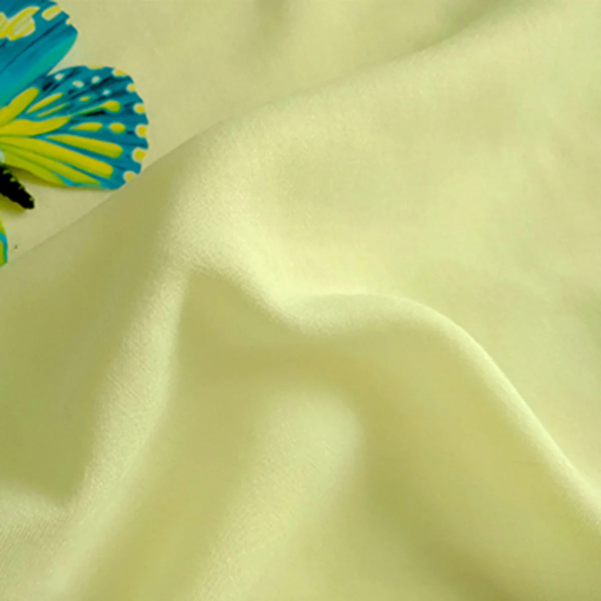 Un gros plan du Tissu jersey viscose uni au mètre de Longan Craft France, 100% viscose rayonne en jaune pâle, montre sa texture lisse et ses plis doux. Un papillon bleu et jaune ajoute du charme à ce tissu en viscose idéal pour créer des robes élégantes.