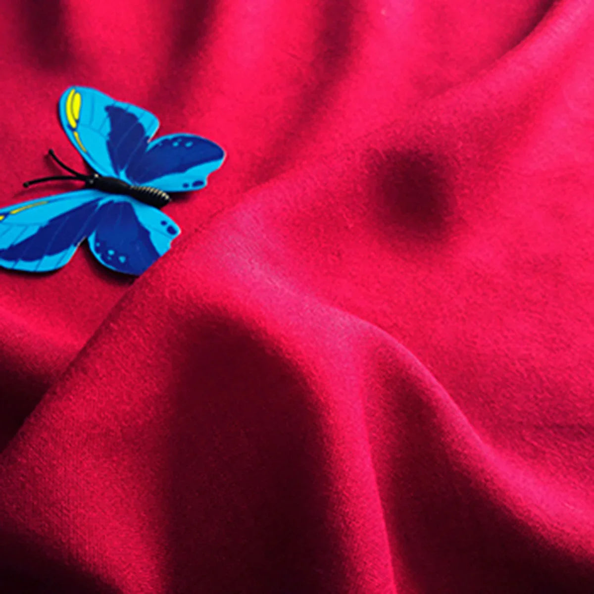 Un papillon bleu se pose sur un tissu jersey viscose uni au mètre, rouge vif et lisse, de Longan Craft France - 100% viscose rayonne, idéal pour les robes, avec des plis souples et des ombres douces.