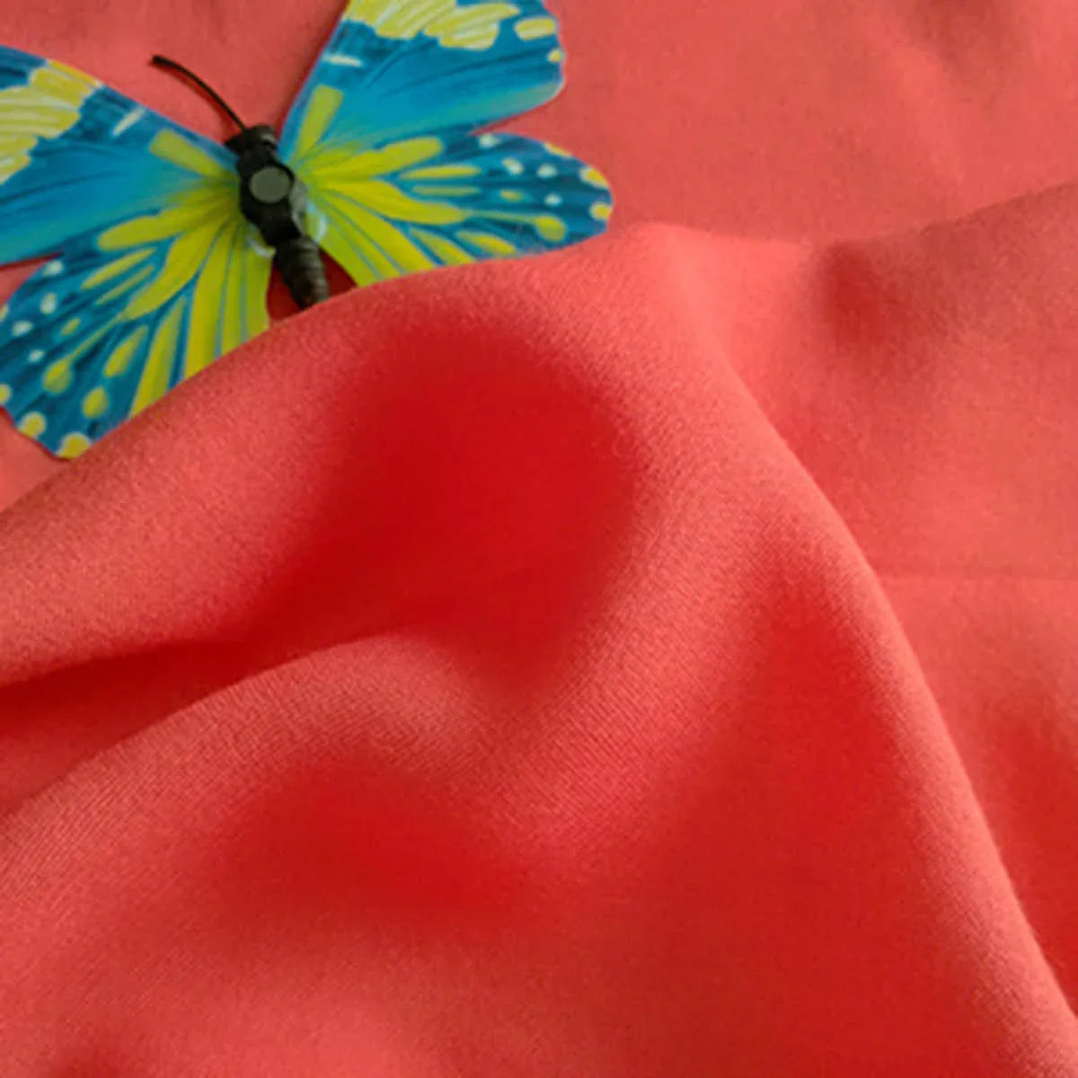 Gros plan sur un Tissu jersey viscose uni au mètre rose corail de Longan Craft France, 100 % viscose rayonne, parfait pour les robes, avec un papillon bleu et jaune sur le dessus ; le tissu lisse présente de légers plis.