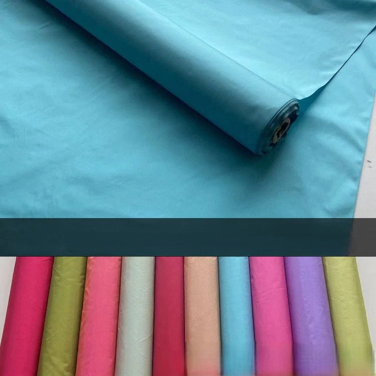 Un rouleau de "Taffetas de soie uni au mètre" turquoise de Longan Craft France est partiellement déroulé, avec des rouleaux verticaux dans des tons vibrants de rose, rouge, vert, bleu et jaune présentés ci-dessous. Fabriqué à 100 % en soie de mûrier, il est idéal pour les robes de mariée.
