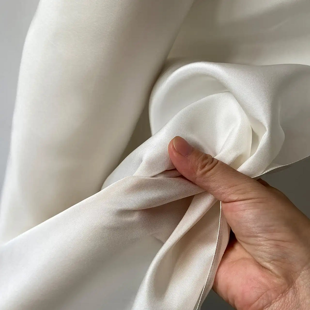 Une main presse délicatement un Taffetas de soie uni au mètre de Longan Craft France-100 % soie de mûrier rigide, un tissu de soie de haute qualité idéal pour les robes de mariée. Le tissu est doux et présente une brillance subtile.