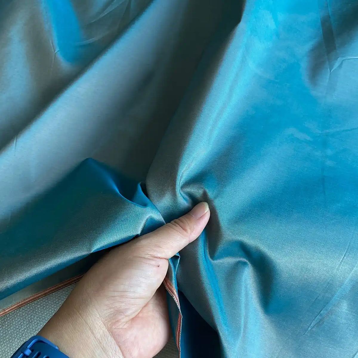 Une main rassemble un morceau de Taffetas de soie uni au mètre de Longan Craft France, 100 % soie de mûrier dans un dégradé de bleu à gris avec des plis visibles. La personne porte un bracelet bleu. Idéal pour les robes de mariée - tissu de soie rigide de haute qualité.