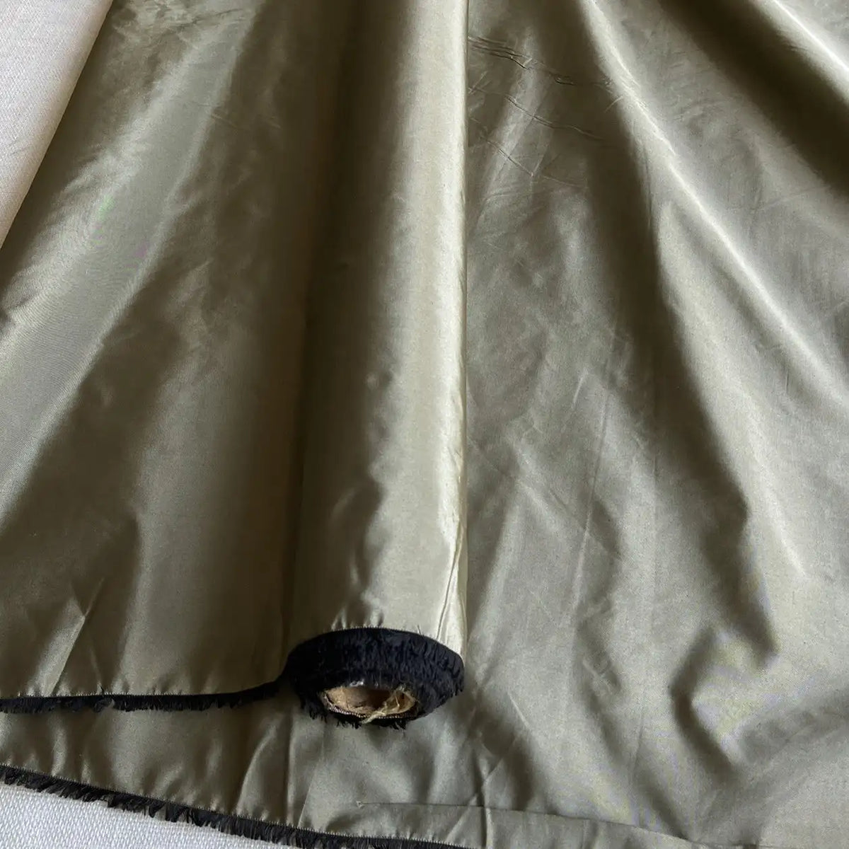 Un morceau de Taffetas de soie uni au mètre de Longan Craft France, 100% soie de mûrier en vert olive clair avec une bordure noire et une petite tache d'usure près du coin, exposant la matière sous-jacente - idéal pour les robes de mariée.