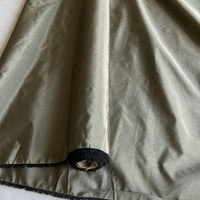 Un morceau de Taffetas de soie uni au mètre de Longan Craft France, 100% soie de mûrier en vert olive clair avec une bordure noire et une petite tache d'usure près du coin, exposant la matière sous-jacente - idéal pour les robes de mariée.