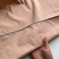 Une main tient un morceau de Taffetas de soie uni au mètre de Longan Craft France, 100 % soie de mûrier en pêche claire avec une couture visible et une bande transparente cousue le long du bord - idéal pour les robes de mariée.