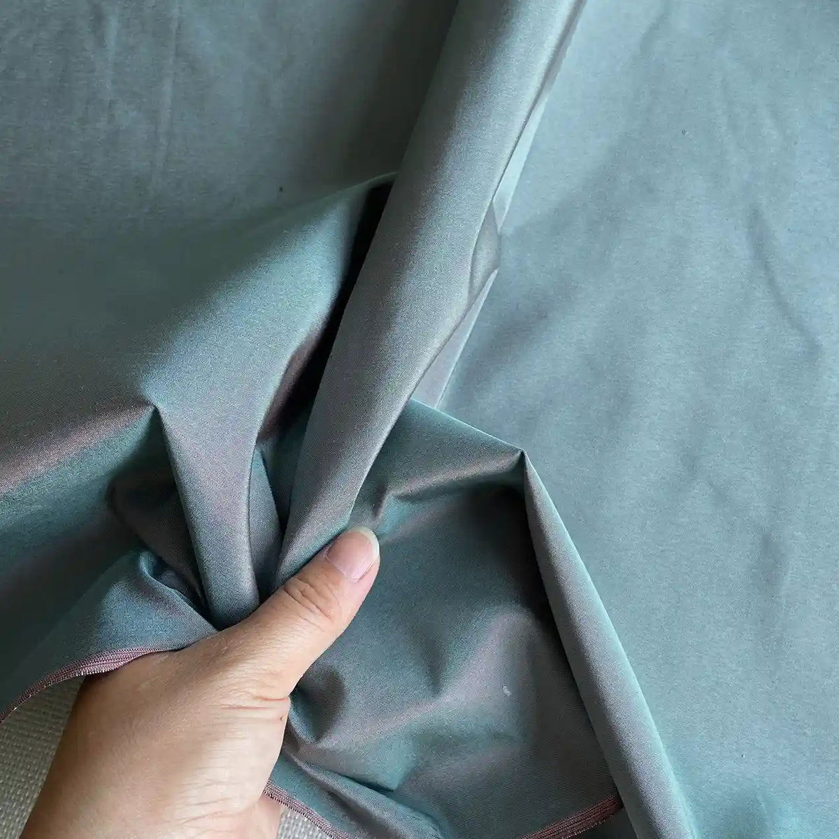 Une main soulève une section du Taffetas de soie uni au mètre de Longan Craft France, un tissu 100% soie de mûrier bleu-gris, lisse et brillant, avec une touche de rose le long du bord, idéal pour les robes de mariée.