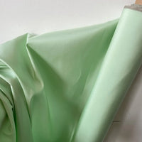 Un rouleau de Taffetas de soie uni au mètre de Longan Craft France, 100 % soie de mûrier en vert clair, est posé contre un mur blanc. Une partie du tissu de haute qualité est drapée pour mettre en valeur sa texture lisse et son éclat subtil, parfaits pour les robes de mariée.