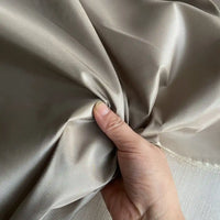 Une main tient un morceau lisse et brillant de Taffetas de soie uni au mètre gris clair de Longan Craft France - un tissu 100 % soie de mûrier, parfait pour les robes de mariée - et l'enroule délicatement sur un fond neutre.