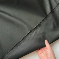 Une main tient le bord du taffetas de soie uni au mètre de Longan Craft France, 100 % soie de mûrier. Ce tissu rigide de haute qualité, destiné aux robes de mariée, est légèrement brillant et laisse apparaître sa couture inachevée sur l'envers.