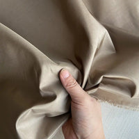Une main tient et forme un léger paquet avec le Taffetas de soie uni au mètre de Longan Craft France, un tissu 100 % soie de mûrier. La surface lisse et brillante reflète la lumière, les bords de ce luxueux tissu de qualité nuptiale étant visibles dans l'angle.