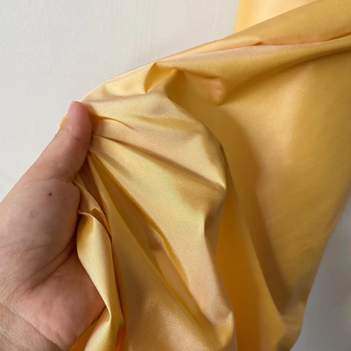 Sur un fond blanc uni, une main rassemble des Taffetas de soie uni au mètre de Longan Craft France (100 % soie de mûrier), brillants et jaunes, parfaits pour les robes de mariée.