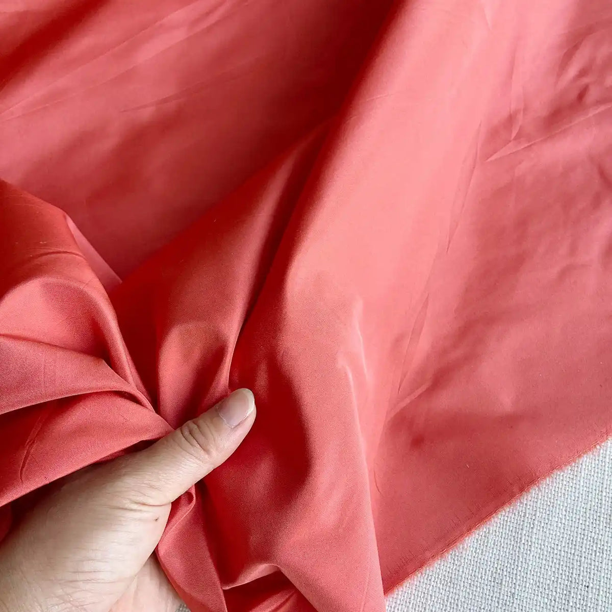 Une main rassemble un morceau de Taffetas de soie uni au mètre de Longan Craft France, un tissu 100 % soie de mûrier destiné aux robes de mariée, mettant en valeur sa texture lisse et rigide et sa brillance rose corail sur un fond clair.