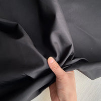Une main pince un morceau de Taffetas de soie uni au mètre noir et lisse de Longan Craft France - un tissu 100 % soie de mûrier parfait pour les robes de mariée - sur une surface claire.