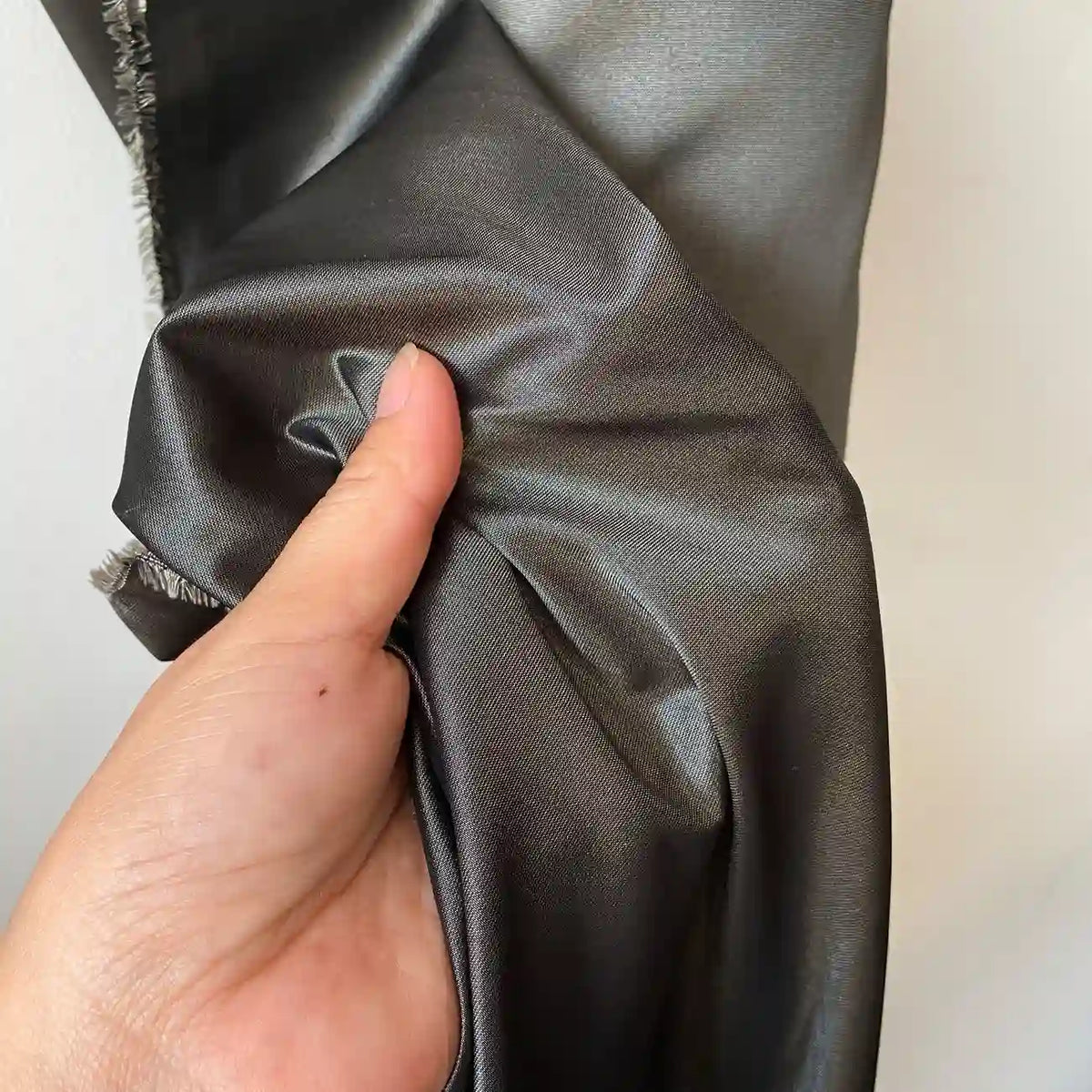 Une main présente un morceau de Taffetas de soie uni au mètre de Longan Craft France, un tissu 100% soie de mûrier gris foncé et brillant, à la texture lisse et réfléchissante, idéal pour les robes de mariée de haute qualité.