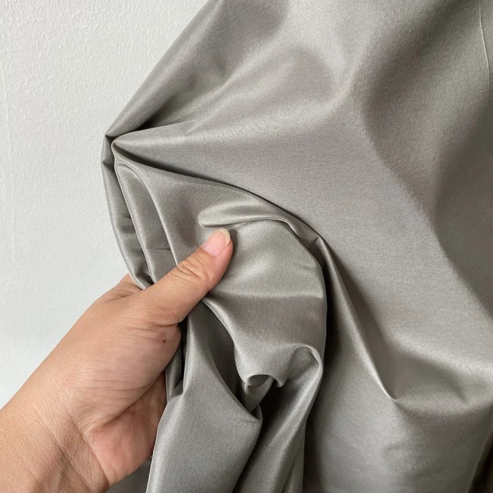 Une main tient un morceau de Longan Craft France Taffetas de soie uni au mètre, tissu 100% soie de mûrier rigide pour robes de mariée, montrant sa texture lisse et gris clair sur un fond blanc uni.