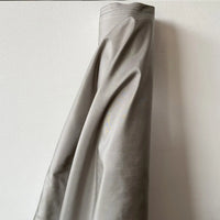 Un rouleau de taffetas de soie uni au mètre de Longan Craft France, 100 % soie de mûrier pour robes de mariée, se tient droit contre un mur blanc. La soie de haute qualité présente une texture lisse, une légère brillance et des plis visibles.