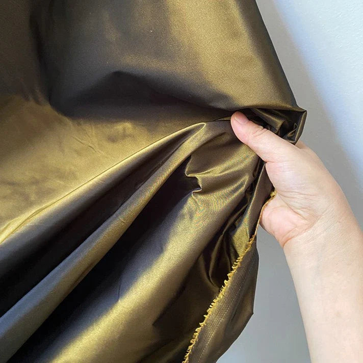 Une main présente un morceau de Longan Craft France Taffetas de soie uni au mètre - tissu en soie de mûrier à 100 %, rigide avec une surface dorée lisse et légèrement réfléchissante et un bord brut visible - idéal pour les robes de mariée de haute qualité.