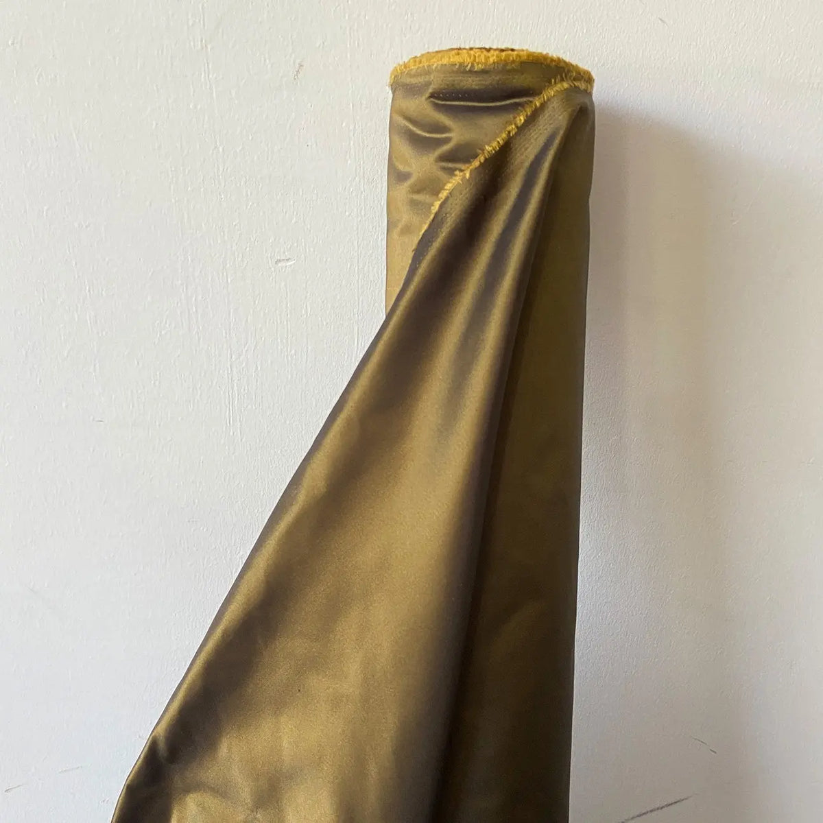 Un rouleau de Longan Craft France Taffetas de soie uni au mètre, 100 % soie de mûrier pour robes de mariée, à l'ourlet jauni, est adossé à un mur blanc uni.