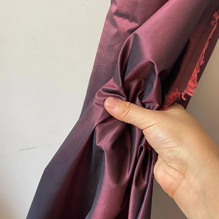 Sur un mur clair, une main saisit un morceau de Longan Craft France Taffetas de soie uni au mètre - 100 % de soie de mûrier rigide, légèrement froissée avec des bords effilochés - idéal pour les robes de mariée de haute qualité.