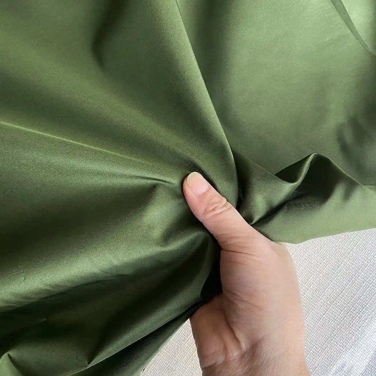 Une main saisit un brillant Taffetas de soie uni au mètre vert olive de Longan Craft France, composé à 100 % de soie de mûrier. La texture lisse et la brillance subtile de ce tissu renforcent son attrait luxueux, idéal pour les robes de mariée de grande qualité.