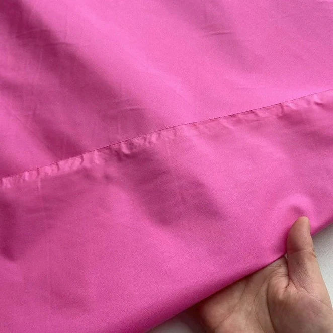 Une main présente des Taffetas de soie uni au mètre rose vif de Longan Craft France, mettant en valeur le tissu lisse 100 % soie de mûrier et un ourlet fini. L'arrière-plan est uni et de couleur claire. Parfait pour les robes de mariée de haute qualité.