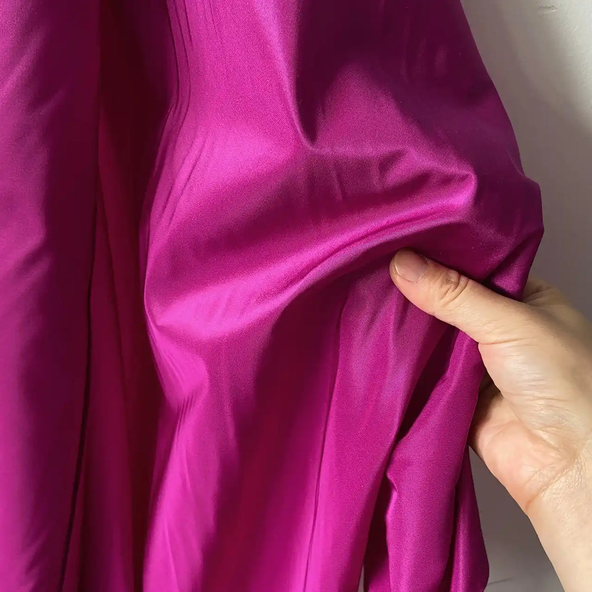Une main tient un morceau de Taffetas de soie uni au mètre de Longan Craft France, un tissu 100 % soie de mûrier, contre un mur clair pour mettre en valeur sa texture lisse et brillante, parfaite pour les robes de mariée de haute qualité.