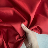 Une main saisit un morceau de Longan Craft France Taffetas de soie uni au mètre, tissu 100% soie de mûrier rigide pour robes de mariée, de couleur rouge vif. La soie de haute qualité présente des plis souples et une légère brillance sur un fond clair.