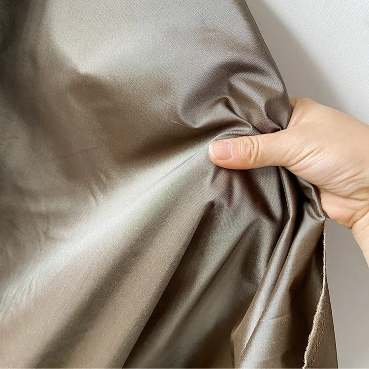 Une main saisit et froisse légèrement le Taffetas de soie uni au mètre de Longan Craft France, un tissu lisse et brillant 100 % soie de mûrier destiné aux robes de mariée, sur un fond clair.