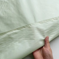 Une main soulève le bord d'une soie lisse et légère, révélant de fines coutures - un échantillon élégant de Taffetas de soie uni au mètre de Longan Craft France, 100 % soie de mûrier, parfait pour les robes de mariée.
