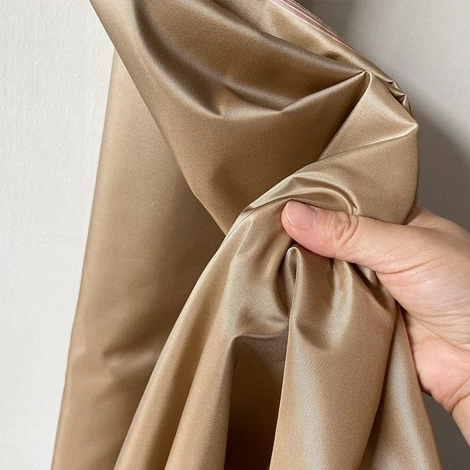 Une main tient un morceau de Longan Craft France Taffetas de soie uni au mètre, 100 % soie de mûrier. Ce tissu brun clair de haute qualité présente des plis lisses et un éclat réfléchissant sur un mur pâle. Parfait pour les robes de mariée.
